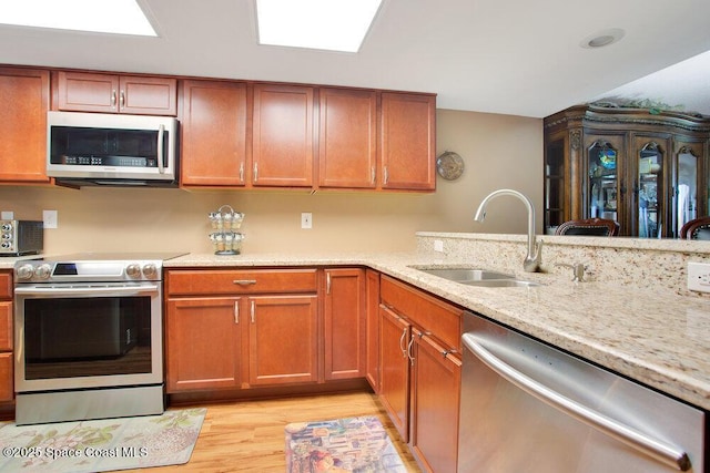 kitchen with brown cabinetry, light stone countertops, a sink, light wood-style flooring, and stainless steel appliances