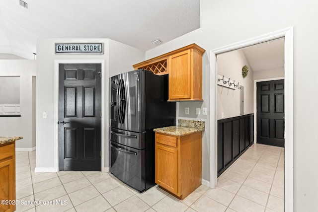kitchen with black fridge with ice dispenser, light tile patterned floors, light stone counters, baseboards, and brown cabinets