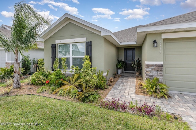 doorway to property featuring a lawn, an attached garage, a shingled roof, stone siding, and stucco siding