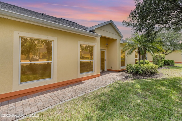 back of house at dusk featuring a shingled roof, a patio area, stucco siding, and a lawn