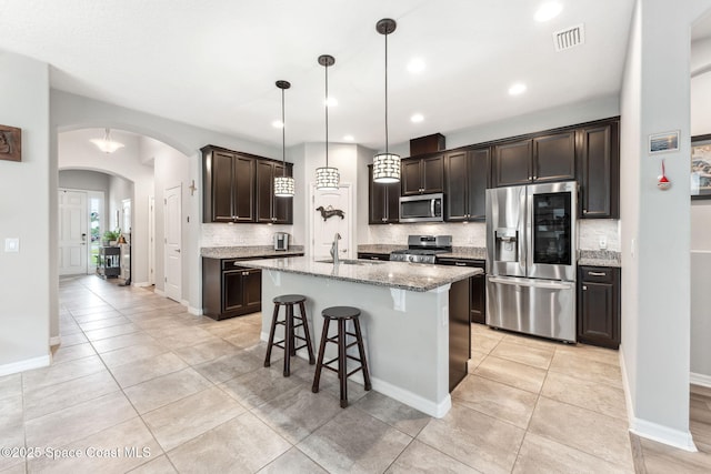 kitchen featuring light stone countertops, dark brown cabinets, tasteful backsplash, arched walkways, and stainless steel appliances
