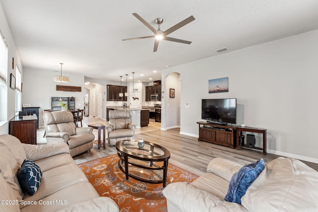 living room with visible vents, light wood-style floors, baseboards, ceiling fan, and arched walkways