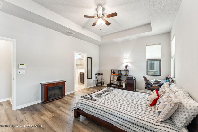 bedroom with visible vents, light wood-style floors, baseboards, a tray ceiling, and ensuite bath