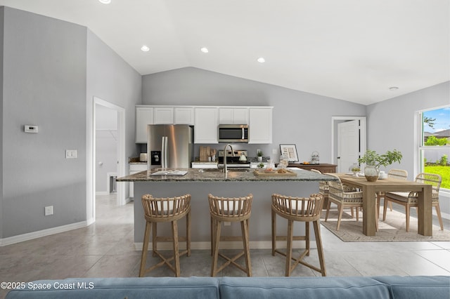 kitchen featuring stainless steel appliances, dark stone countertops, an island with sink, white cabinets, and lofted ceiling