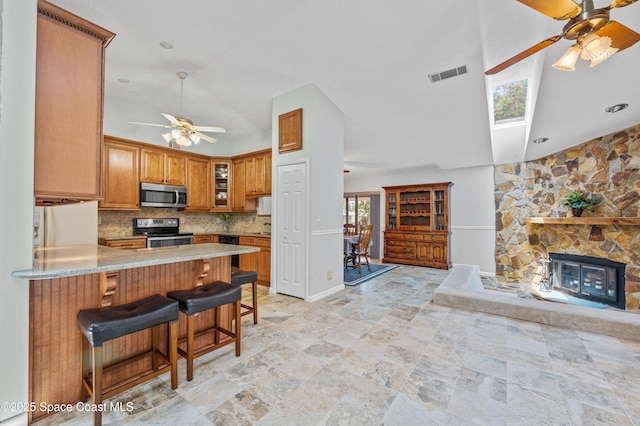 kitchen with appliances with stainless steel finishes, a ceiling fan, decorative backsplash, brown cabinets, and glass insert cabinets