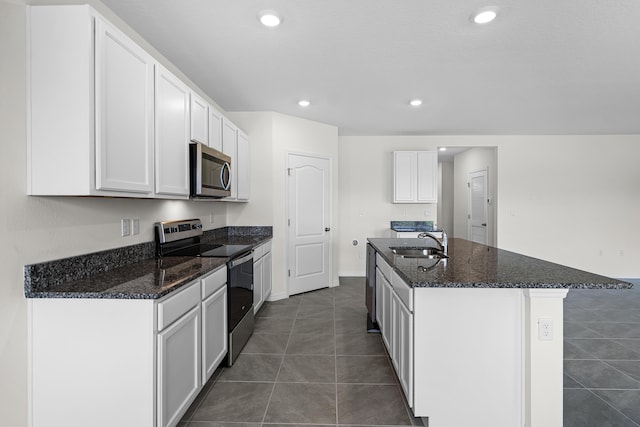 kitchen with a kitchen island with sink, dark tile patterned floors, dark stone counters, a sink, and appliances with stainless steel finishes