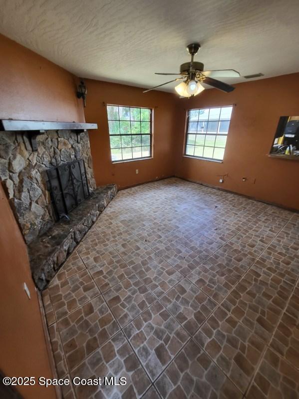 unfurnished living room with a textured ceiling, a stone fireplace, stone finish floor, a ceiling fan, and visible vents