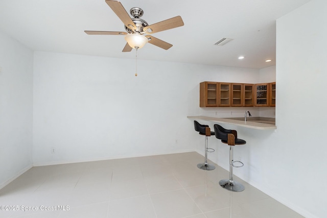 kitchen with brown cabinets, a kitchen bar, glass insert cabinets, light countertops, and ceiling fan