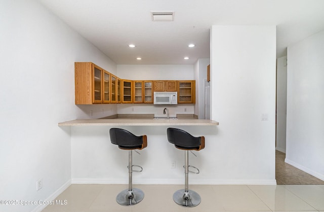kitchen featuring white microwave, brown cabinetry, a sink, glass insert cabinets, and a kitchen bar