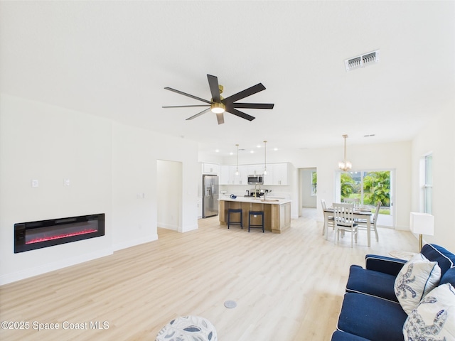 living area with light wood-style flooring, ceiling fan with notable chandelier, visible vents, baseboards, and a glass covered fireplace