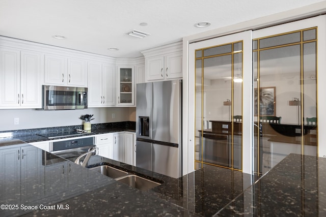 kitchen with a sink, dark stone countertops, white cabinetry, glass insert cabinets, and appliances with stainless steel finishes