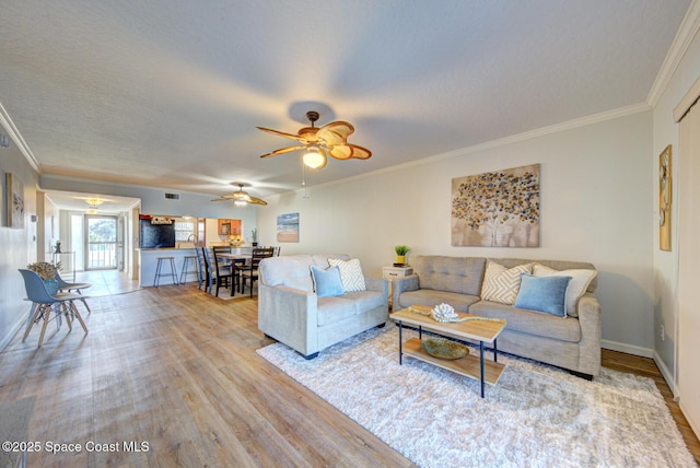 living area with wood finished floors, ornamental molding, ceiling fan, a textured ceiling, and baseboards