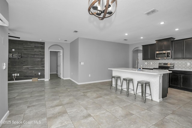 kitchen with arched walkways, appliances with stainless steel finishes, a kitchen island with sink, recessed lighting, and baseboards