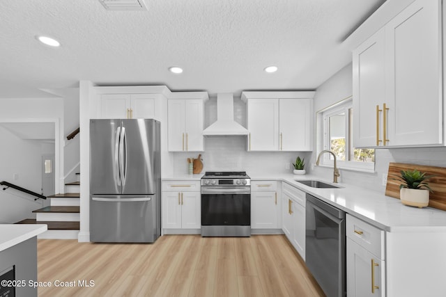 kitchen featuring appliances with stainless steel finishes, wall chimney exhaust hood, a sink, light wood-type flooring, and a textured ceiling