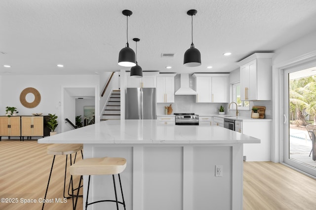 kitchen featuring appliances with stainless steel finishes, wall chimney range hood, light wood finished floors, a sink, and a kitchen island