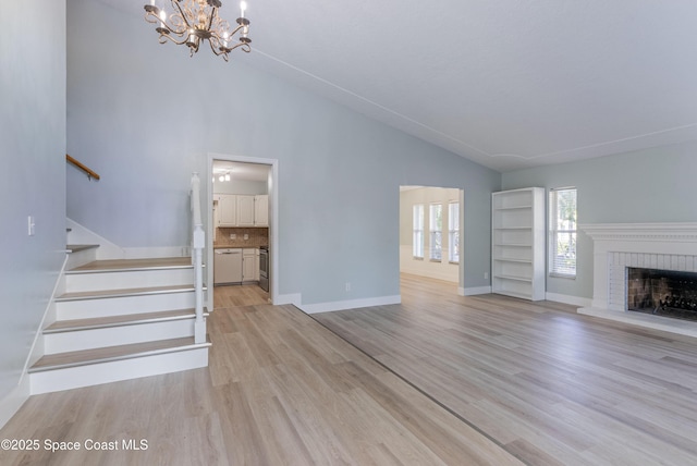 unfurnished living room featuring a chandelier, stairway, light wood-style floors, a brick fireplace, and baseboards