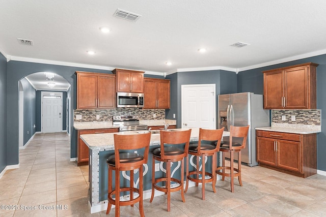 kitchen featuring stainless steel appliances, arched walkways, light countertops, backsplash, and a center island with sink