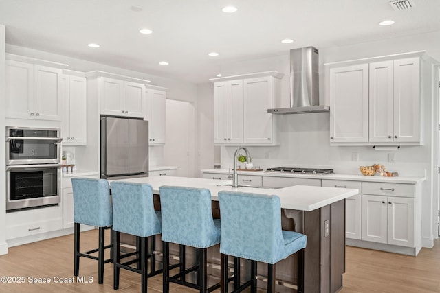 9374 Corvo Ct, Melbourne, FL 32940 - kitchen featuring wall chimney range hood, appliances with stainless steel finishes, a kitchen island with sink, light wood-style flooring, and recessed lighting