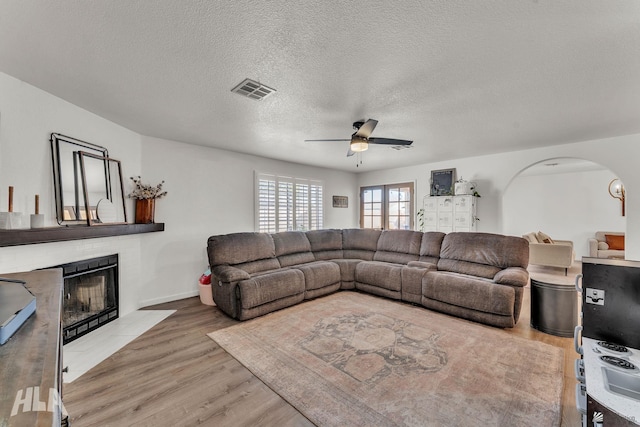 living area with light wood finished floors, arched walkways, a ceiling fan, a fireplace with flush hearth, and a textured ceiling