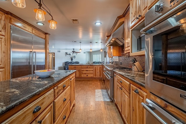 kitchen featuring pendant lighting, high end appliances, wall chimney exhaust hood, tasteful backsplash, and light hardwood / wood-style floors