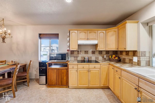 kitchen featuring black electric stovetop, tile counters, sink, light brown cabinetry, and a notable chandelier