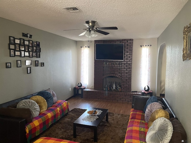 living room featuring wood-type flooring, a brick fireplace, a textured ceiling, and ceiling fan