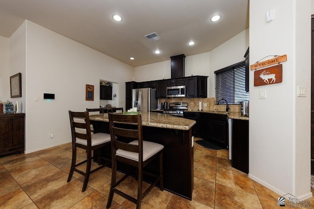 kitchen featuring a kitchen bar, appliances with stainless steel finishes, a kitchen island, light stone countertops, and decorative backsplash