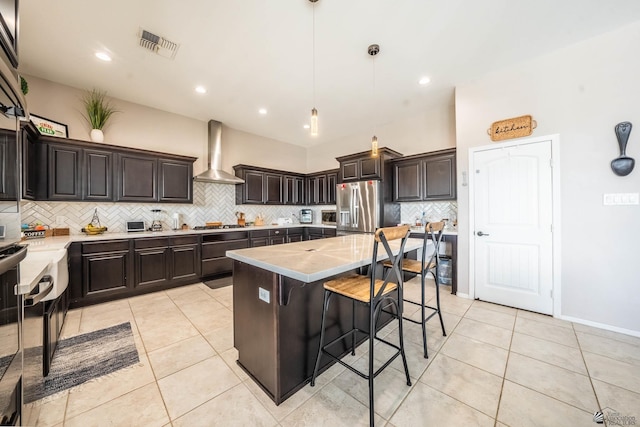 kitchen with wall chimney range hood, stainless steel fridge, a center island, dark brown cabinetry, and decorative light fixtures