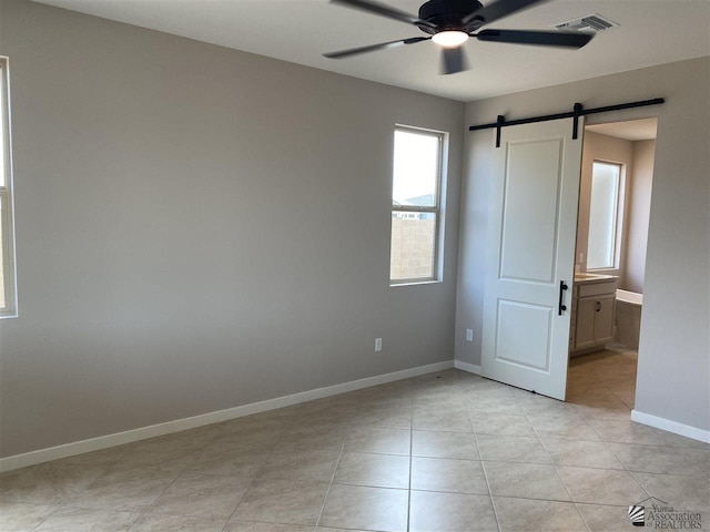 unfurnished bedroom featuring ceiling fan, a barn door, light tile patterned flooring, and ensuite bathroom