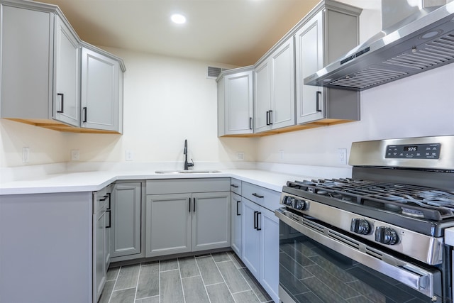 kitchen featuring wall chimney range hood, stainless steel gas stove, light countertops, gray cabinetry, and a sink