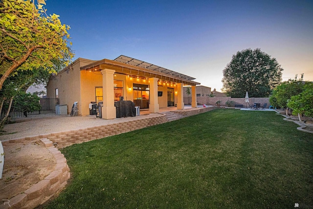 back of property at dusk with a fenced backyard, a patio, stucco siding, and a yard