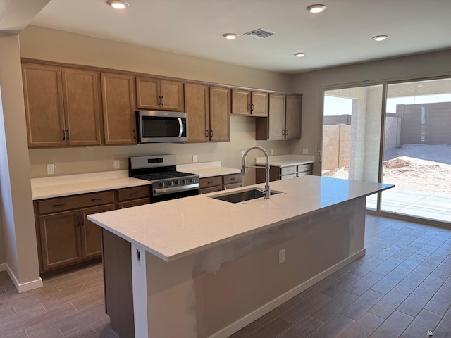 kitchen with wood finish floors, appliances with stainless steel finishes, a sink, light countertops, and an island with sink