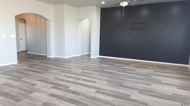 unfurnished room featuring a ceiling fan, arched walkways, wood finished floors, baseboards, and a decorative wall