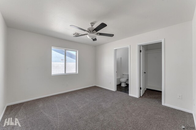 unfurnished bedroom with a walk in closet, ceiling fan, baseboards, ensuite bath, and dark colored carpet