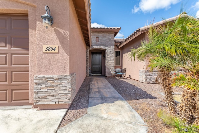 doorway to property with stucco siding, stone siding, a garage, and a tiled roof