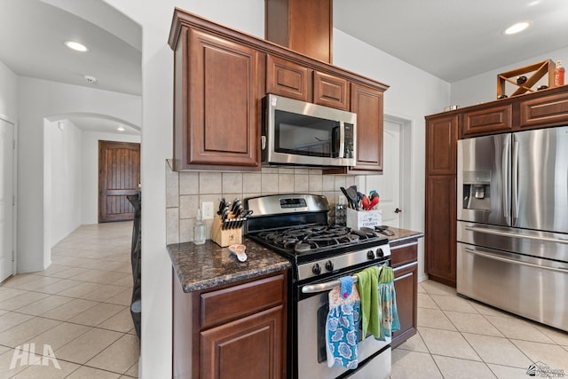 kitchen featuring backsplash, arched walkways, dark stone countertops, stainless steel appliances, and light tile patterned flooring