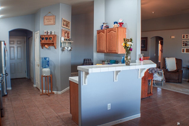 kitchen with arched walkways, tile counters, a peninsula, baseboards, and a kitchen bar