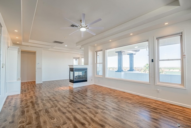 unfurnished living room with visible vents, wood finished floors, ceiling fan, a tray ceiling, and a multi sided fireplace