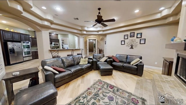 living room featuring a fireplace, ceiling fan, recessed lighting, a raised ceiling, and arched walkways