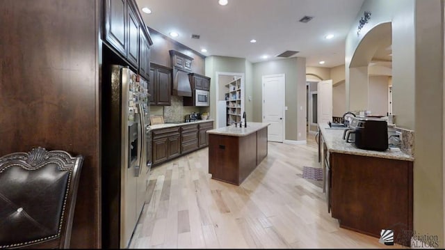 kitchen featuring dark brown cabinetry, stainless steel refrigerator with ice dispenser, arched walkways, light wood-style floors, and an island with sink