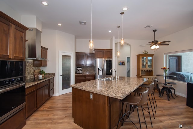 kitchen featuring wall chimney range hood, light stone counters, backsplash, an island with sink, and black appliances