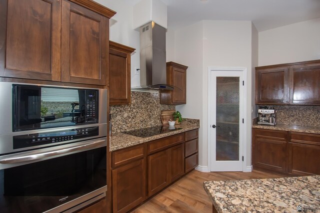 kitchen with black appliances, wall chimney range hood, decorative backsplash, light stone countertops, and light wood-type flooring