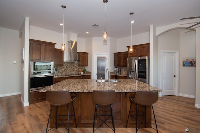 kitchen with sink, wall chimney exhaust hood, hanging light fixtures, a large island with sink, and appliances with stainless steel finishes