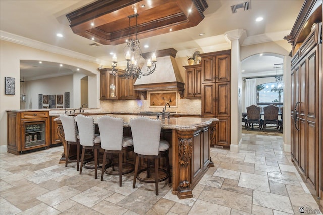 kitchen with light stone counters, kitchen peninsula, an island with sink, a tray ceiling, and custom exhaust hood