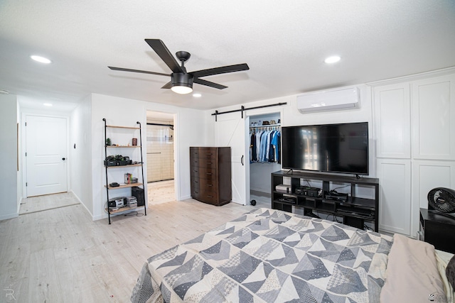 bedroom with a wall mounted air conditioner, light wood-type flooring, ceiling fan, a barn door, and a closet