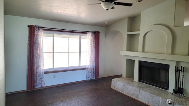 unfurnished living room featuring ceiling fan, a tiled fireplace, a textured ceiling, built in shelves, and dark hardwood / wood-style flooring