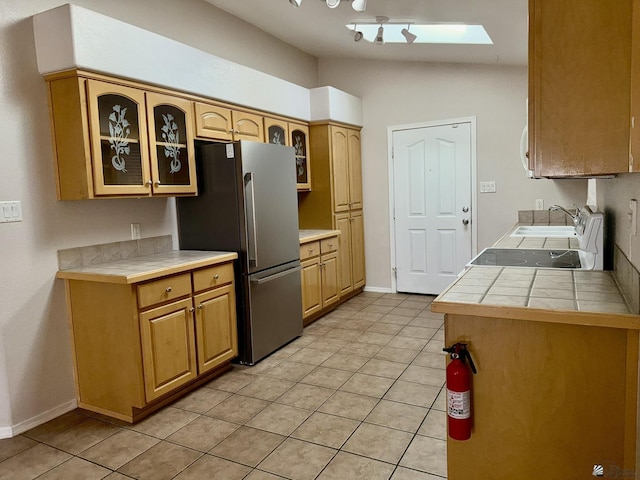 kitchen featuring tile countertops, stainless steel fridge, vaulted ceiling with skylight, light tile patterned flooring, and sink