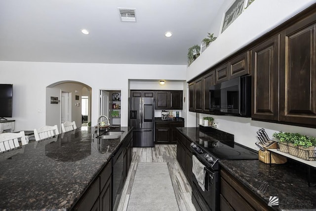 kitchen featuring sink, a breakfast bar area, dark brown cabinets, dark stone counters, and black appliances