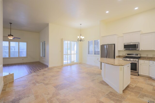 kitchen with ceiling fan with notable chandelier, appliances with stainless steel finishes, a kitchen island, light stone counters, and white cabinetry