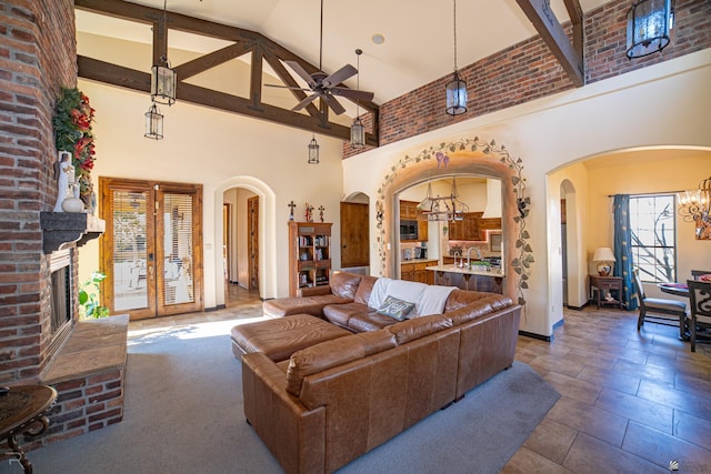 living room with a brick fireplace, ceiling fan with notable chandelier, sink, beam ceiling, and high vaulted ceiling